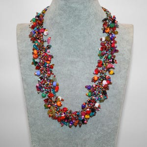 May include: A multi-strand statement necklace featuring a dense cluster of small, colorful glass beads and larger irregular stone-like beads in shades of red, orange, yellow, green, blue, and purple, creating a textured, vibrant, and layered appearance.