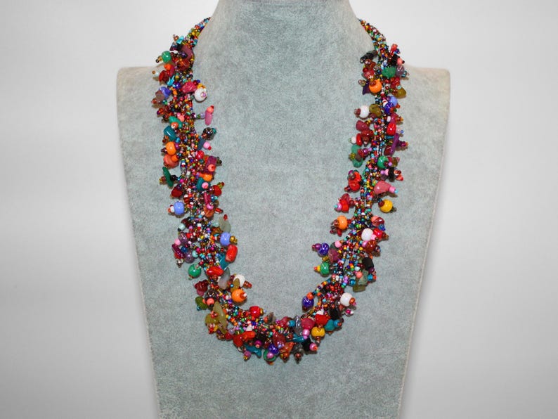 May include: A multi-strand statement necklace featuring a dense cluster of small, colorful glass beads and larger irregular stone-like beads in shades of red, orange, yellow, green, blue, and purple, creating a textured, vibrant, and layered appearance.