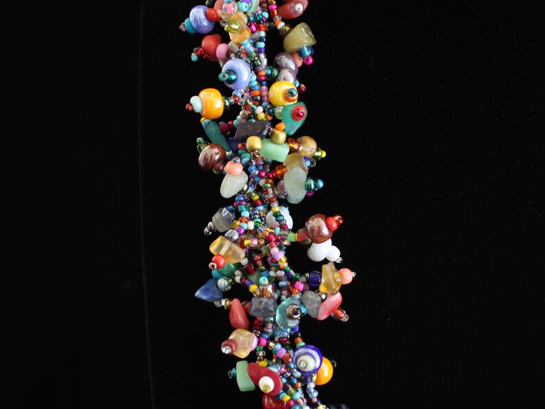May include: A multi-strand cluster necklace featuring an assortment of colorful beads in various shapes and sizes. The design incorporates small seed beads, larger smooth round beads, and irregular stone-like pieces in shades of red, blue, yellow, green, and purple.