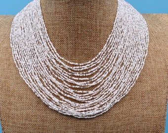 May include: A white beaded necklace with multiple strands of small, round beads. The necklace is displayed on a brown burlap mannequin.
