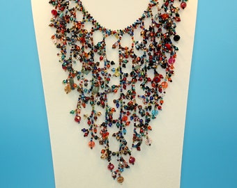 May include: A colorful beaded necklace with a cascading design. The necklace features a variety of beads in shades of red, orange, yellow, green, blue, and purple. The beads are arranged in a delicate pattern that creates a flowing, waterfall effect.