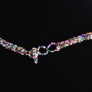 May include: A multi-colored beaded necklace featuring a twisted rope design made from tiny glass seed beads in various vibrant hues. The necklace includes a circular beaded clasp mechanism and additional decorative bead clusters at the ends.