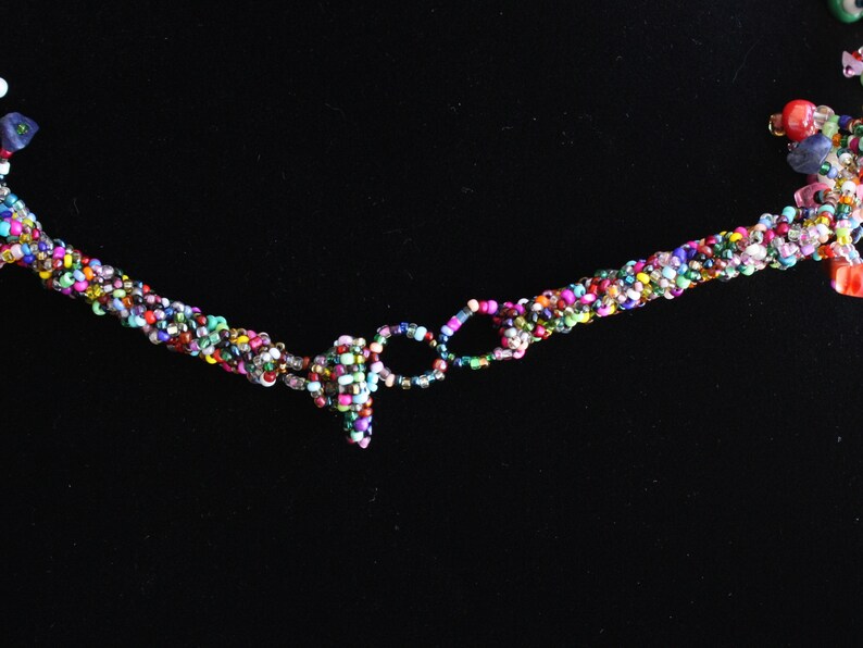 May include: A multi-colored beaded necklace featuring a twisted rope design made from tiny glass seed beads in various vibrant hues. The necklace includes a circular beaded clasp mechanism and additional decorative bead clusters at the ends.