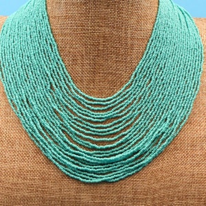 May include: A turquoise blue beaded necklace with multiple strands. The necklace is displayed on a brown burlap mannequin.