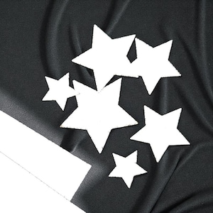 Puede incluir: Un fondo de tela negra con varias formas de estrellas blancas dispersas. Las estrellas varían en tamaño y están recortadas con un aspecto ligeramente texturizado. Una forma rectangular blanca está en la esquina inferior izquierda.