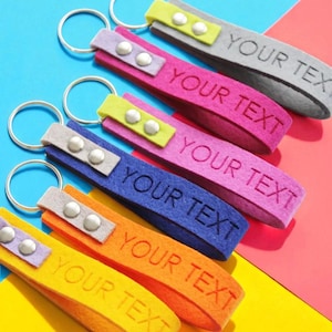 Op de afbeelding: Een verzameling kleurrijke sleutelhangers met de tekst "YOUR TEXT" in reliëf op elke band. De sleutelhangers hebben verschillende kleuren, waaronder geel, oranje, blauw, roze en grijs, met zilveren metalen sleutelringen en decoratieve accenten.