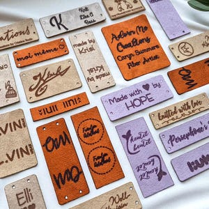 May include: A collection of rectangular, laser-engraved tags in various colors, including tan, orange, and lavender. The tags feature text such as "Criations," "Made with HOPE," and "knitted with love." Some tags have small holes for attaching to items.