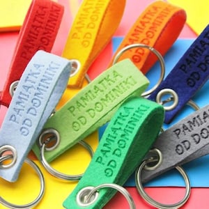Könnte beinhalten: Eine Auswahl an farbenfrohen Schlüsselanhängern in Pink, Rot, Gelb, Orange, Grün, Blau und Grau. Jeder Schlüsselanhänger hat eine Stoffschlaufe mit dem Text "PAMIĄTKA OD DOMINIKI" und einen silberfarbenen Metallring.