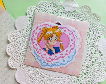 Puede incluir: Una pegatina troquelada que muestra a un personaje de anime rubio con dos moños y cuello azul, enmarcado por un corazón rosa con un borde blanco similar al encaje. La pegatina está sobre una tarjeta cuadrada a cuadros rosa claro y un tapete.