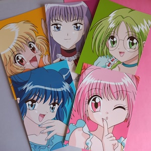 Op de afbeelding: Vijf kleurrijke anime-personage trading cards met vrouwelijke personages met verschillende haarkleuren en -stijlen. De personages zijn getekend in een cartoon-stijl met grote ogen en felle kleuren.