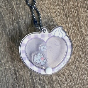 May include: A clear, heart-shaped keychain with a silver ball chain. The keychain features a light purple gingham pattern and a cartoon character. The character is holding a lollipop. The keychain has the text "Pok&eacute;mon (TM) TOMY".