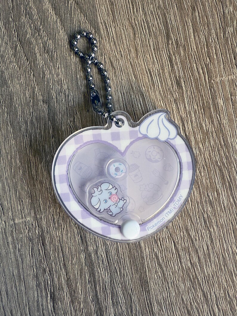 May include: A clear, heart-shaped keychain with a silver ball chain. The keychain features a light purple gingham pattern and a cartoon character. The character is holding a lollipop. The keychain has the text "Pok&eacute;mon (TM) TOMY".