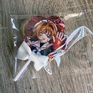 May include: A clear plastic package containing a heart-shaped anime character keychain with a white bow on a stick. The keychain features a character with orange hair, a red and white outfit, and a waving hand.