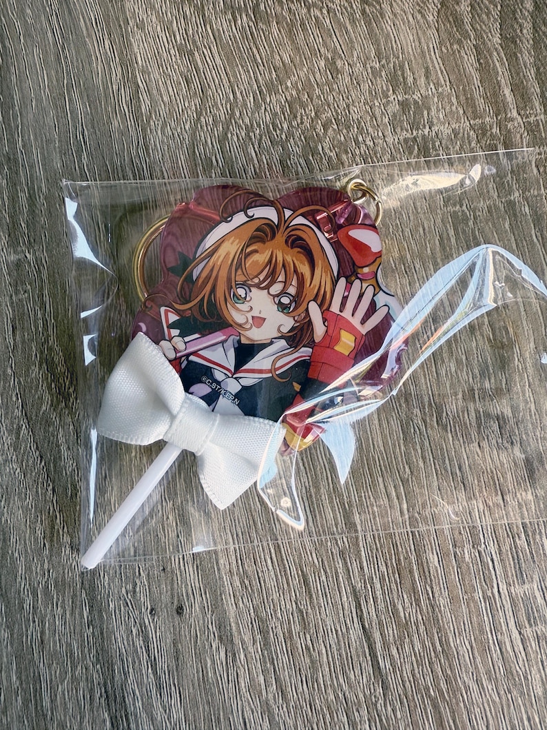 May include: A clear plastic package containing a heart-shaped anime character keychain with a white bow on a stick. The keychain features a character with orange hair, a red and white outfit, and a waving hand.
