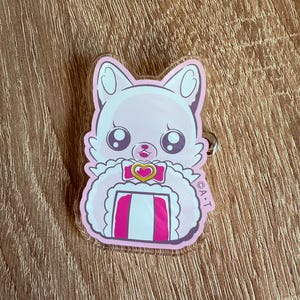 May include: A pink and white cartoon cat keychain with large eyes and a heart-shaped bow. The cat has a fluffy collar and a striped design on its chest. The keychain is made of clear acrylic and has a silver-colored ring.