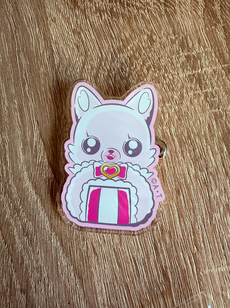 May include: A pink and white cartoon cat keychain with large eyes and a heart-shaped bow. The cat has a fluffy collar and a striped design on its chest. The keychain is made of clear acrylic and has a silver-colored ring.