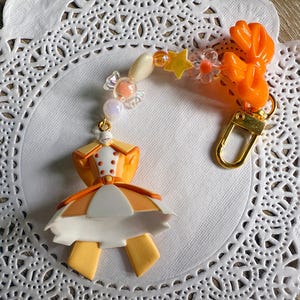 May include: A handmade charm featuring a miniature orange and white dress with a gold-colored clasp. The charm is adorned with beads, stars, and flower accents. The charm is attached to a gold-colored clasp.