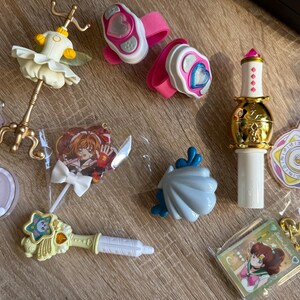 May include: A collection of colorful anime-themed toys and accessories. Includes a golden wand, pink wrist cuffs with heart designs, a ballerina figurine, and character keychains. The items are in various shapes and sizes.