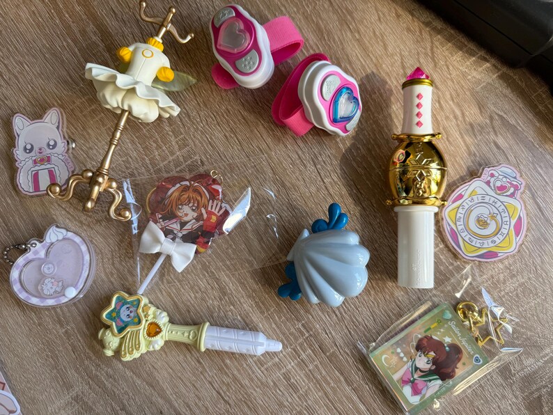 May include: A collection of colorful anime-themed toys and accessories. Includes a golden wand, pink wrist cuffs with heart designs, a ballerina figurine, and character keychains. The items are in various shapes and sizes.