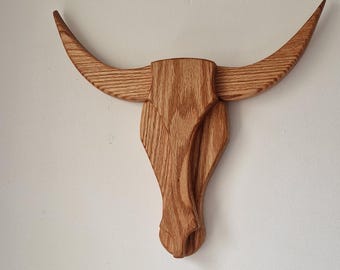 May include: Wooden bull head wall decor. The sculpture is made of light brown wood and features a detailed carving of a bull's head with curved horns. A rustic accent for home or office.