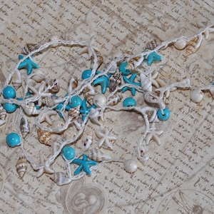 Può includere: Una collana di corda bianca con conchiglie e stelle marine blu e bianche. La collana è su una superficie con motivo bianco e marrone.