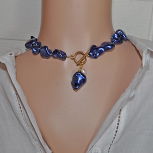 Op de afbeelding: Een blauwe statement ketting met onregelmatig gevormde kralen en een goudkleurige sluiting. Een enkele blauwe hanger met een witte parel hangt aan de sluiting. De ketting wordt op een mannequin getoond.