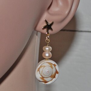Könnte beinhalten: Goldfarbene Ohrstecker mit Seestern-Motiv und hängendem Design. Die Ohrringe haben zwei kleine, hellrosa Perlen und eine große, weiß-braune Muschel. Die Ohrringe sind an einem Ohr abgebildet.