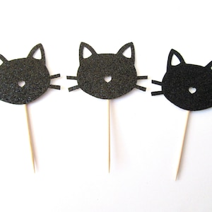 Puede incluir: Tres toppers de cupcake con forma de cabeza de gato de purpurina negra con un dise&ntilde;o sencillo. Cada topper est&aacute; unido a un palo de madera.