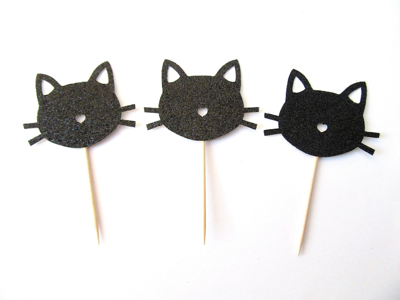 Puede incluir: Tres toppers de cupcake con forma de cabeza de gato de purpurina negra con un dise&ntilde;o sencillo. Cada topper est&aacute; unido a un palo de madera.