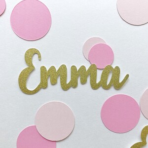 K&ouml;nnte beinhalten: Goldener Glitzerkonfetti mit dem Namen "Emma" in goldenen Glitzerbuchstaben auf wei&szlig;em Hintergrund mit rosa Konfetti-Kreisen.