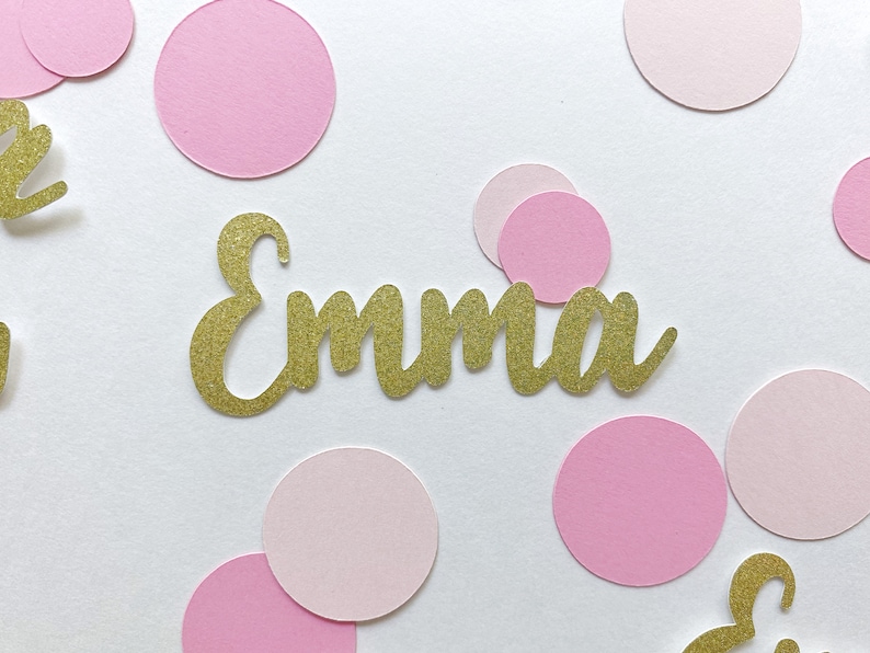 K&ouml;nnte beinhalten: Goldener Glitzerkonfetti mit dem Namen "Emma" in goldenen Glitzerbuchstaben auf wei&szlig;em Hintergrund mit rosa Konfetti-Kreisen.