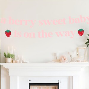 May include: A pink banner with the text "A berry sweet baby is on the way" and two strawberry illustrations. The banner is hanging on a white wall above a white mantelpiece, with decorative items and plants.