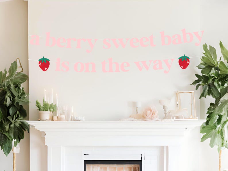 May include: A pink banner with the text "A berry sweet baby is on the way" and two strawberry illustrations. The banner is hanging on a white wall above a white mantelpiece, with decorative items and plants.