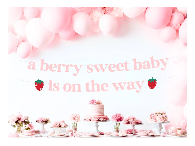 May include: A pink banner with the text "a berry sweet baby is on the way" is displayed against a white background. The banner is decorated with two strawberry illustrations. Pink balloons and floral arrangements complete the baby shower theme.