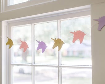 Può includere: Una ghirlanda di unicorni con teste di unicorno dorate, rosa e viola appese a una cornice di finestra bianca.