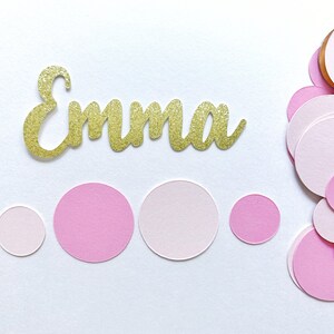 K&ouml;nnte beinhalten: Ein goldener Glitzer-Namens-Ausschnitt mit der Aufschrift "Emma" mit verstreuten rosa Konfetti-Kreisen.