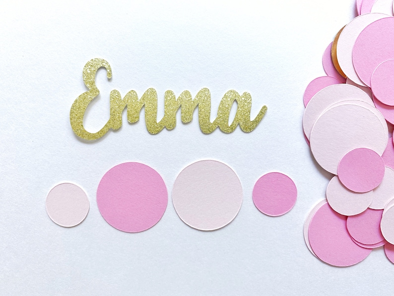 K&ouml;nnte beinhalten: Ein goldener Glitzer-Namens-Ausschnitt mit der Aufschrift "Emma" mit verstreuten rosa Konfetti-Kreisen.