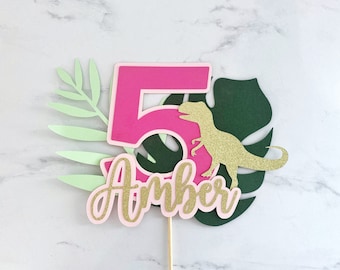 May include: Pink and gold cake topper with a number 5, the name Amber, a green leaf, and a gold glitter dinosaur.