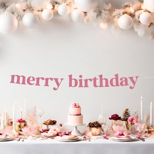 May include: A pink glitter banner with the text "merry birthday" hanging above a table set for a party. The table is decorated with white and pink flowers, candles, and a two-tiered pink cake.