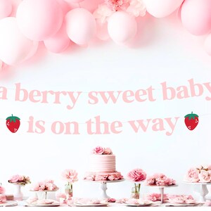 May include: A pink banner with the text "a berry sweet baby is on the way" and strawberry illustrations. The banner is suspended above a table with pink flowers, cakes, and balloons, creating a festive atmosphere for a baby shower.