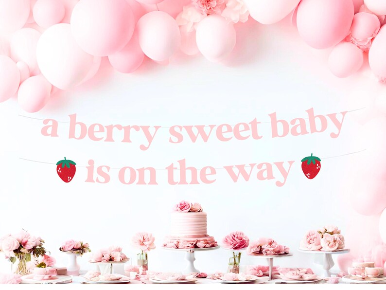 May include: A pink banner with the text "a berry sweet baby is on the way" and strawberry illustrations. The banner is suspended above a table with pink flowers, cakes, and balloons, creating a festive atmosphere for a baby shower.