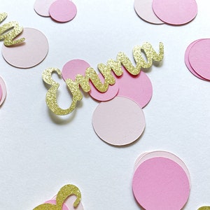 K&ouml;nnte beinhalten: Rosa und goldene Konfetti mit dem Namen "Emma" in goldenen Glitzer geschrieben. Das Konfetti ist auf einem wei&szlig;en Hintergrund verstreut.