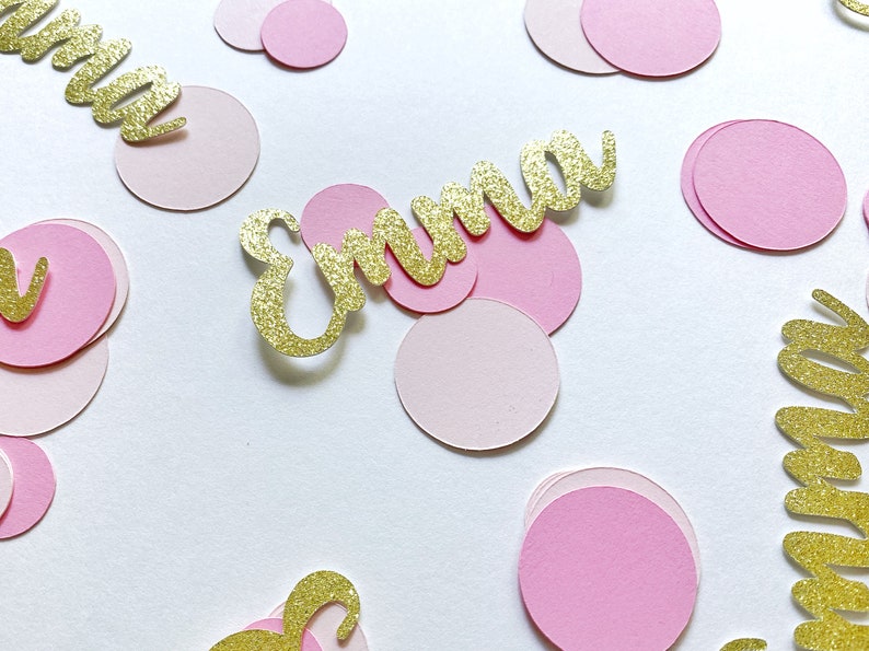 K&ouml;nnte beinhalten: Rosa und goldene Konfetti mit dem Namen "Emma" in goldenen Glitzer geschrieben. Das Konfetti ist auf einem wei&szlig;en Hintergrund verstreut.