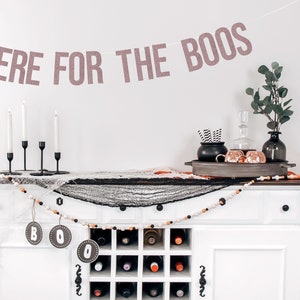 Puede incluir: Un armario blanco con una estantería para vinos y una guirnalda de cuentas blancas y negras con la palabra "BOO" en ella. El armario está decorado con una telaraña negra y una pancarta que dice "HERE FOR THE BOOS".