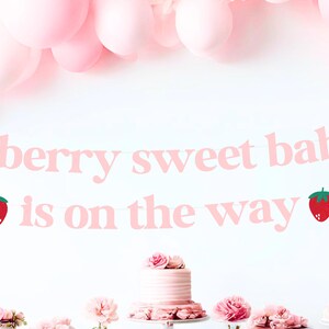 May include: A pink banner with the text "a berry sweet baby is on the way" and strawberry illustrations. Pink balloons are in the background, with a cake and flowers on a table below.