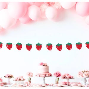 May include: A festive party scene with a strawberry banner strung across the top. Pink balloons and a table with a cake and floral arrangements create a celebratory atmosphere. The banner features red strawberries with green leaves.