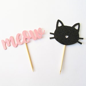 Puede incluir: Dos adornos para cupcakes de papel rosa y negro. Uno dice "miau" y el otro es una cara de gato negro.