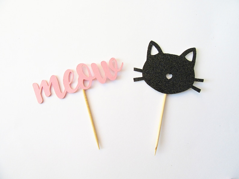 Puede incluir: Dos adornos para cupcakes de papel rosa y negro. Uno dice "miau" y el otro es una cara de gato negro.