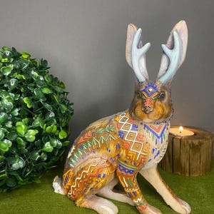 May include: A decorative brown jackalope figurine with blue antlers and colourful geometric patterns. The figurine is sitting on a green surface, with a small candle and a green plant in the background.