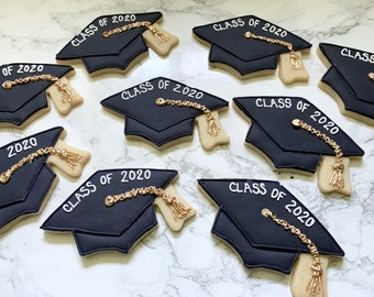 Peut inclure: Biscuits noirs en forme de coiffes de graduation avec l'inscription "CLASS OF 2020" en glaçage blanc. Chaque biscuit est orné d'un gland doré. Les biscuits sont disposés sur une surface en marbre blanc.